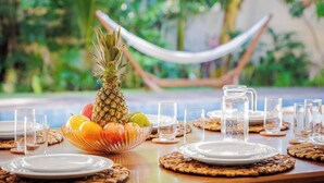 A dining table set with plates, glasses, a pitcher of water, and a bowl of fruit including a pineapple, apples, and oranges. - Deluxe Triple Suite at Villa Lux Do Mar (Jijoca de Jericoacoara)