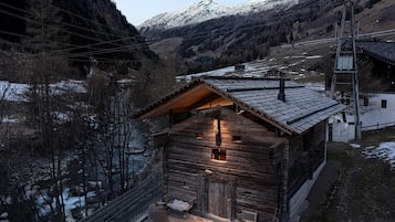 A wooden cabin with a sloped roof, a stone pathway leading to the entrance, and a view of a river and mountains in the background.