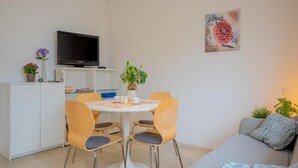 A modern living room with a grey sofa, a dining table with chairs, a flat-screen TV on a stand, and a painting on the wall.