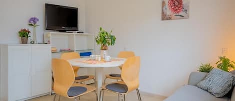 A modern living room with a grey sofa, a dining table with chairs, a flat-screen TV on a stand, and a painting on the wall.