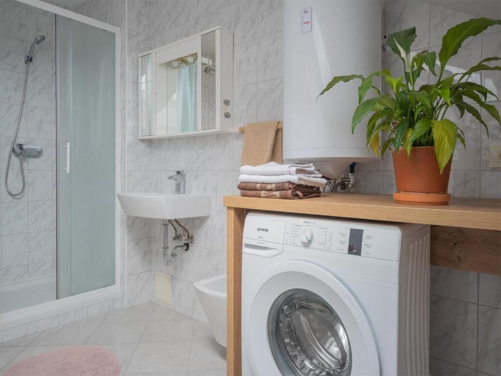A modern bathroom with a glass shower enclosure, a built-in sink, a washing machine, and a potted plant.