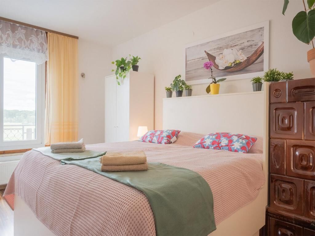 Een slaapkamer met een bed, kussens, een raam met gordijnen, een wandplank met planten en een houten kast.