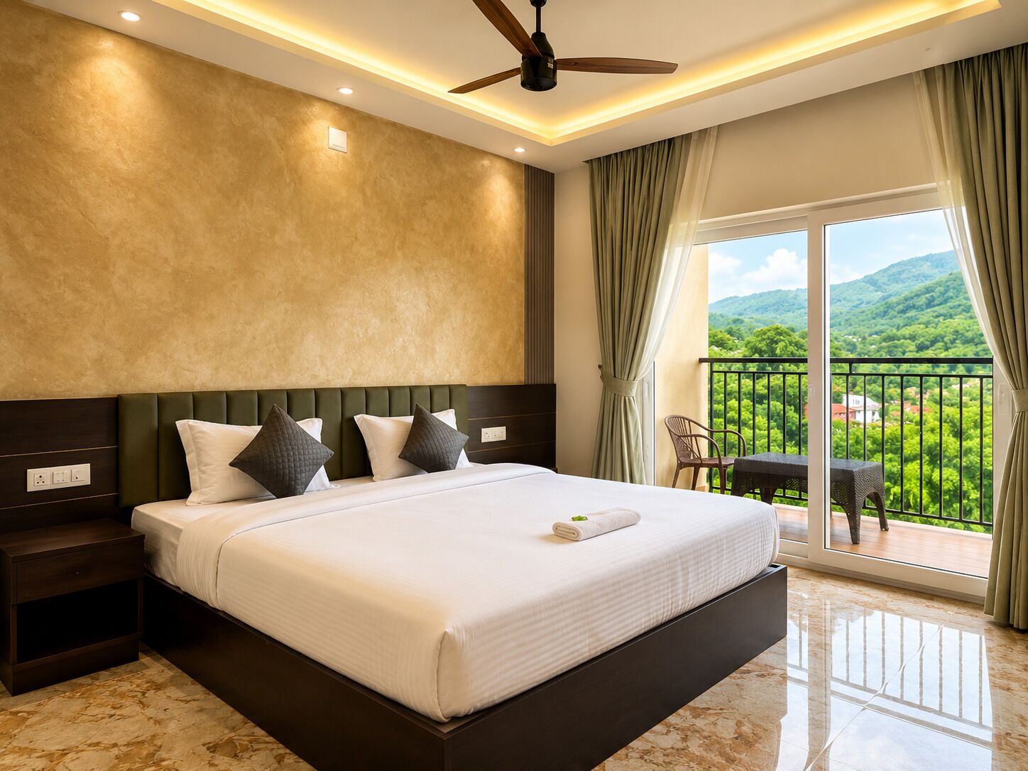 A hotel room with a large bed, a ceiling fan, a view of greenery through the balcony, and a table with chairs outside.