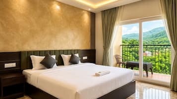 A hotel room with a large bed, a ceiling fan, a view of greenery through the balcony, and a table with chairs outside.