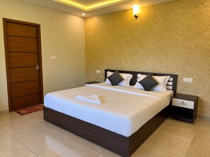 A hotel room with a large bed, a wooden door, a nightstand, and a wall-mounted light fixture. - Foggy Woods Resort Wayanad (Vythiri)