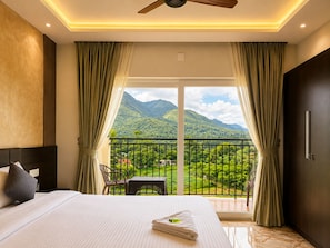 A hotel room with a bed, a table, chairs, a balcony with a view of mountains, and a ceiling fan. - Foggy Woods Resort Wayanad (Vythiri)