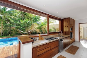 A modern kitchen with a large window, wooden cabinets, a gas stove, and a built-in dishwasher. - Deluxe King Suite at Villa Lux Do Mar (Jijoca de Jericoacoara)