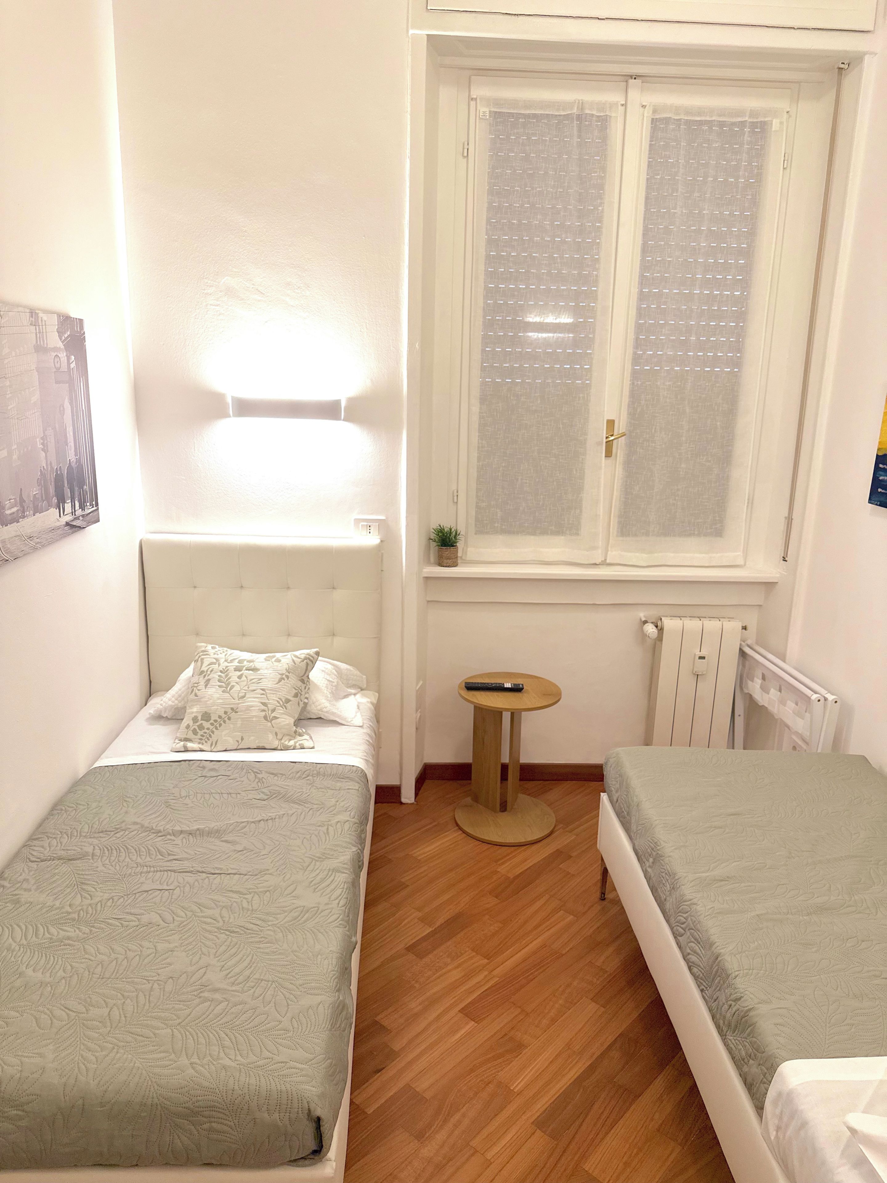 Un dormitorio pequeño y bien iluminado con dos camas individuales, una mesita de noche, un radiador y una ventana con persianas.
