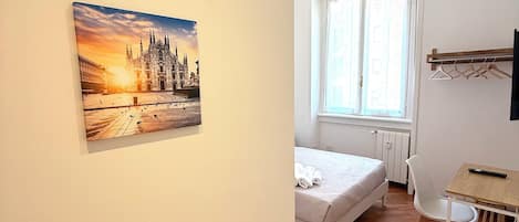 Quarto com cama, escrivaninha, cadeira, janela e um quadro na parede.