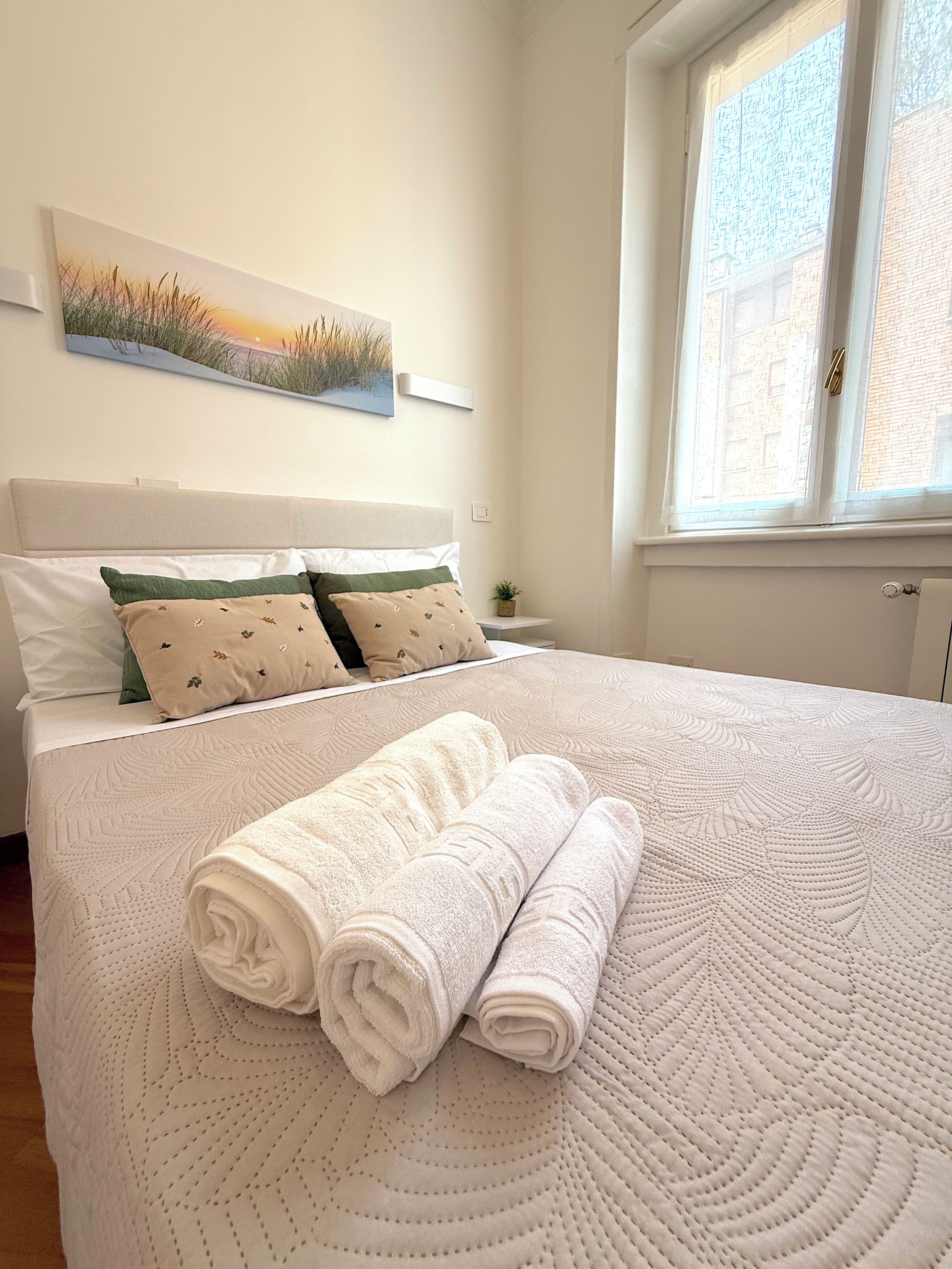 A neatly made bed with rolled white towels, a headboard, and a wall-mounted painting.