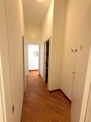 Un pasillo con piso de madera, paredes blancas y una puerta que da a otra habitación.