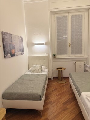 Una habitación pequeña y luminosa con dos camas individuales, una mesa redonda y un ventanal con persianas.