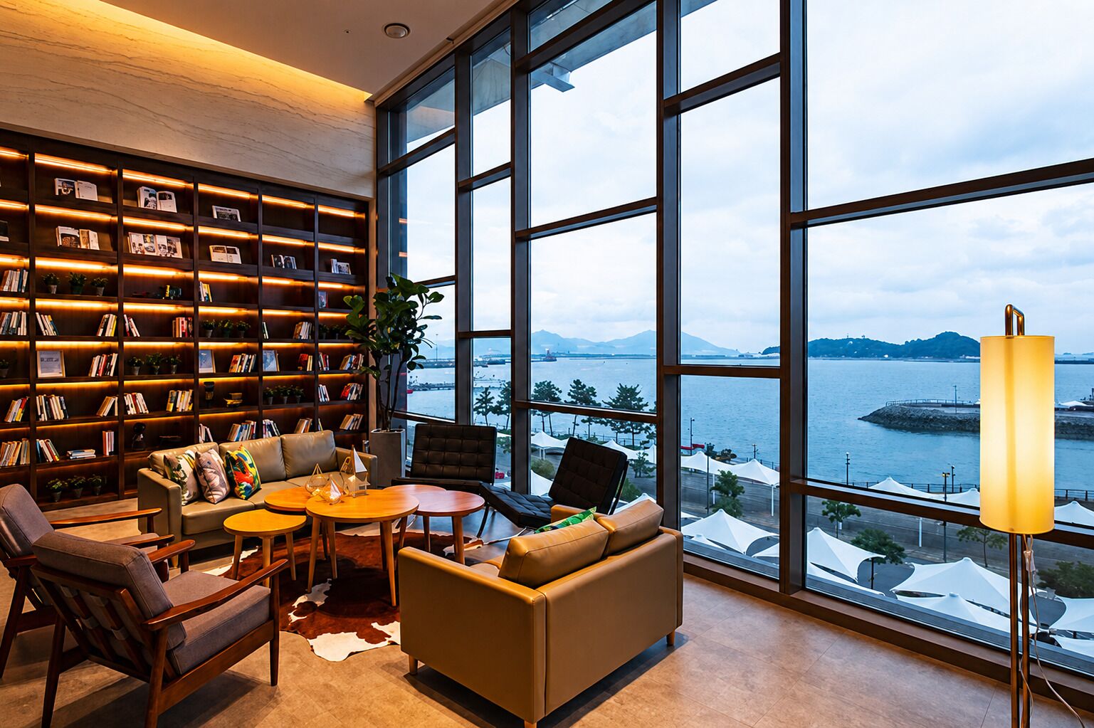 A modern interior with a large window overlooking a body of water, a bookshelf filled with books, and a seating area with a table and chairs.