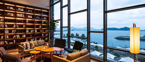 A modern interior with a large window overlooking a body of water, a bookshelf filled with books, and a seating area with a table and chairs.