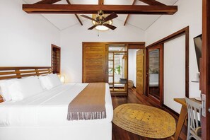 Une chambre avec un grand lit, des poutres apparentes en bois au plafond, un ventilateur de plafond et une vue sur un balcon, accessible par une porte ouverte.