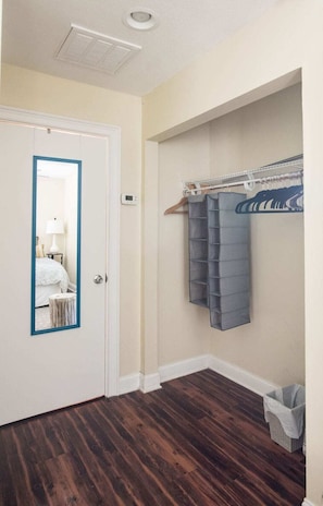 A room with a wooden floor, a white door with a blue frame, a mirror, a wall-mounted drying rack, and a small trash bin. - Cozy Jasmine Cottage | Savannah + Tybee l Cohens Retreat (Savannah)