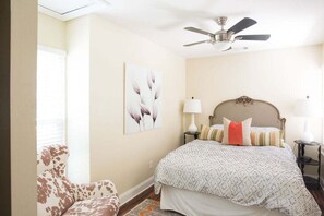 A bedroom with a bed, a ceiling fan, a lamp, a chair, and a wall art piece. - Cozy Jasmine Cottage | Savannah + Tybee l Cohens Retreat (Savannah)