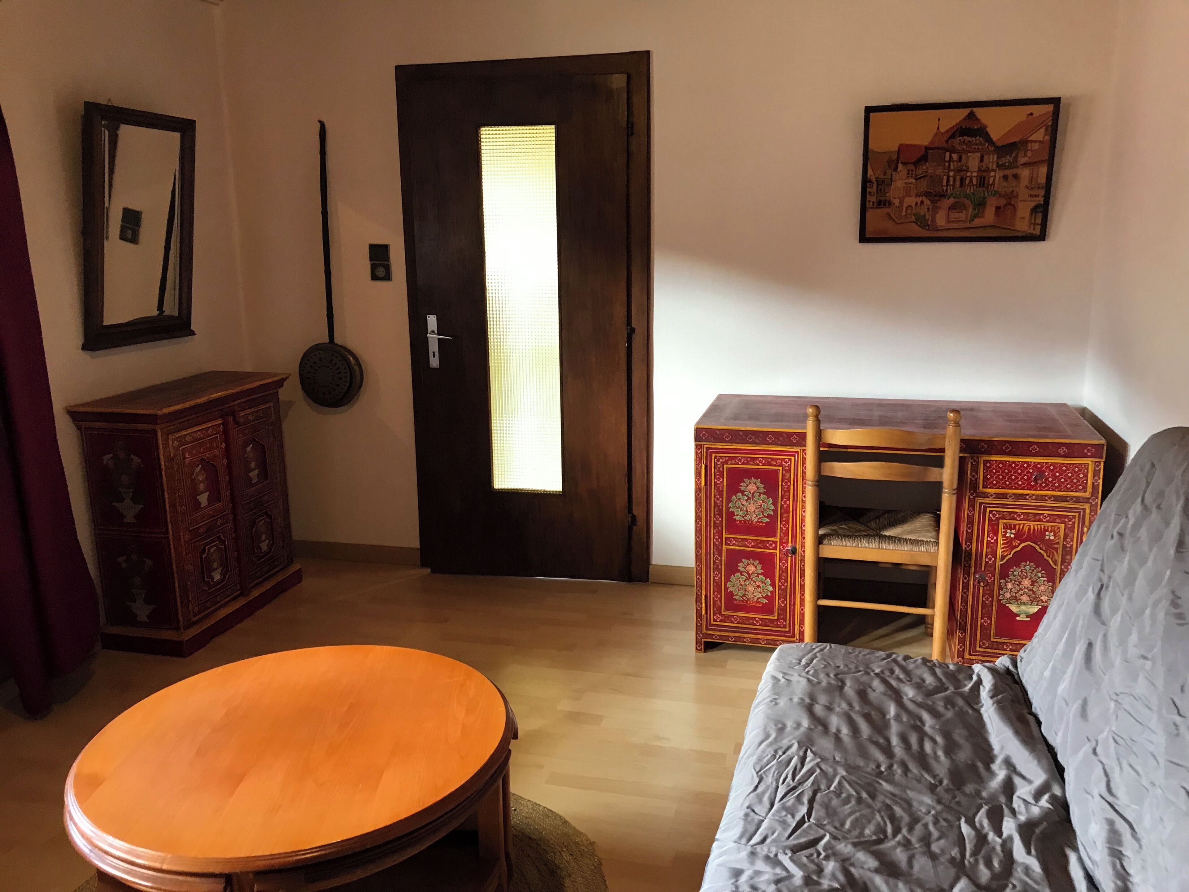 Een kamer met een houten deur, een ronde tafel, een bank, een kast en een aan de muur hangend schilderij.