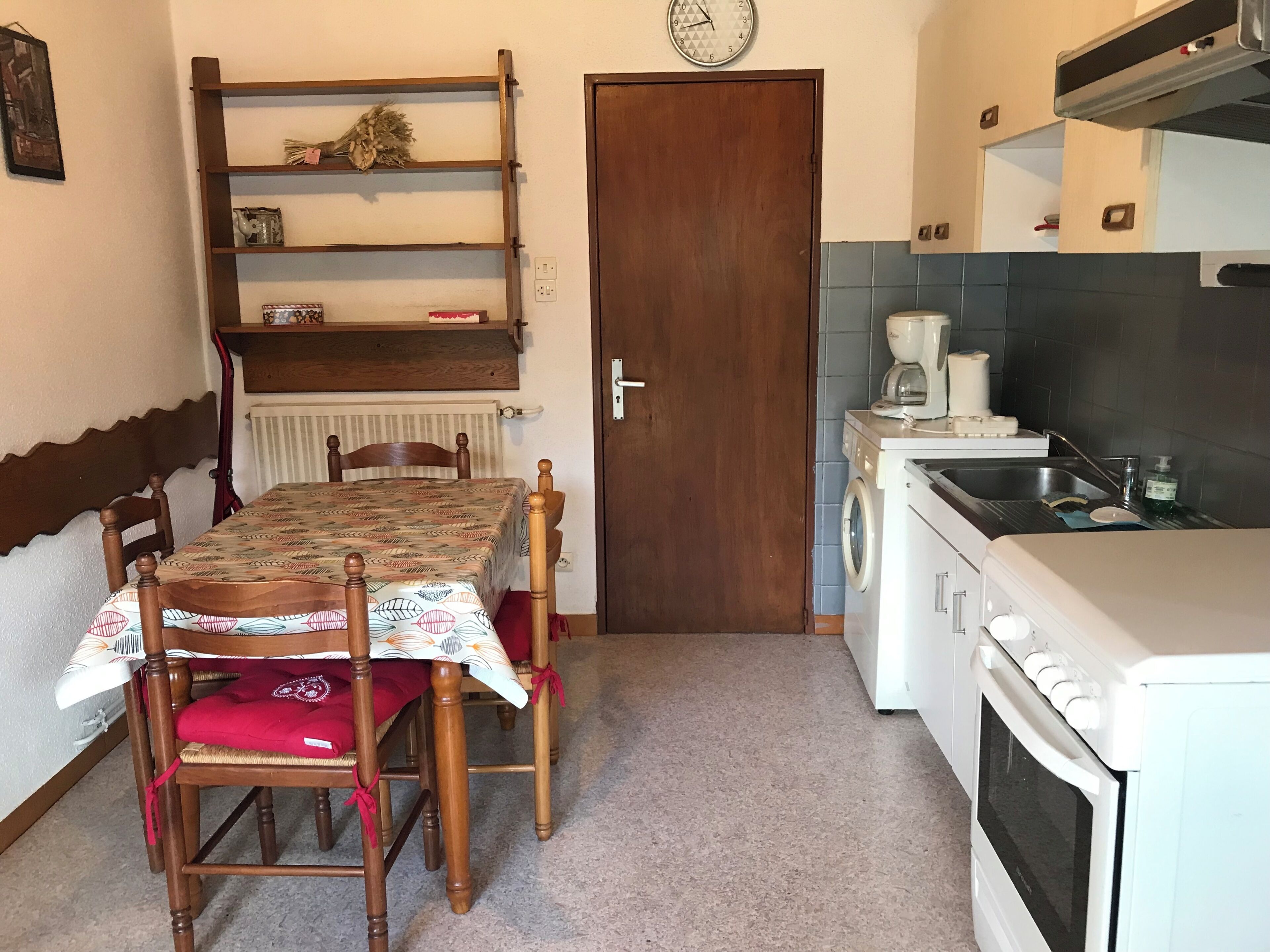 Een kleine keuken met een eettafel, stoelen, een koffiezetapparaat, een wasmachine en een klok aan de muur.