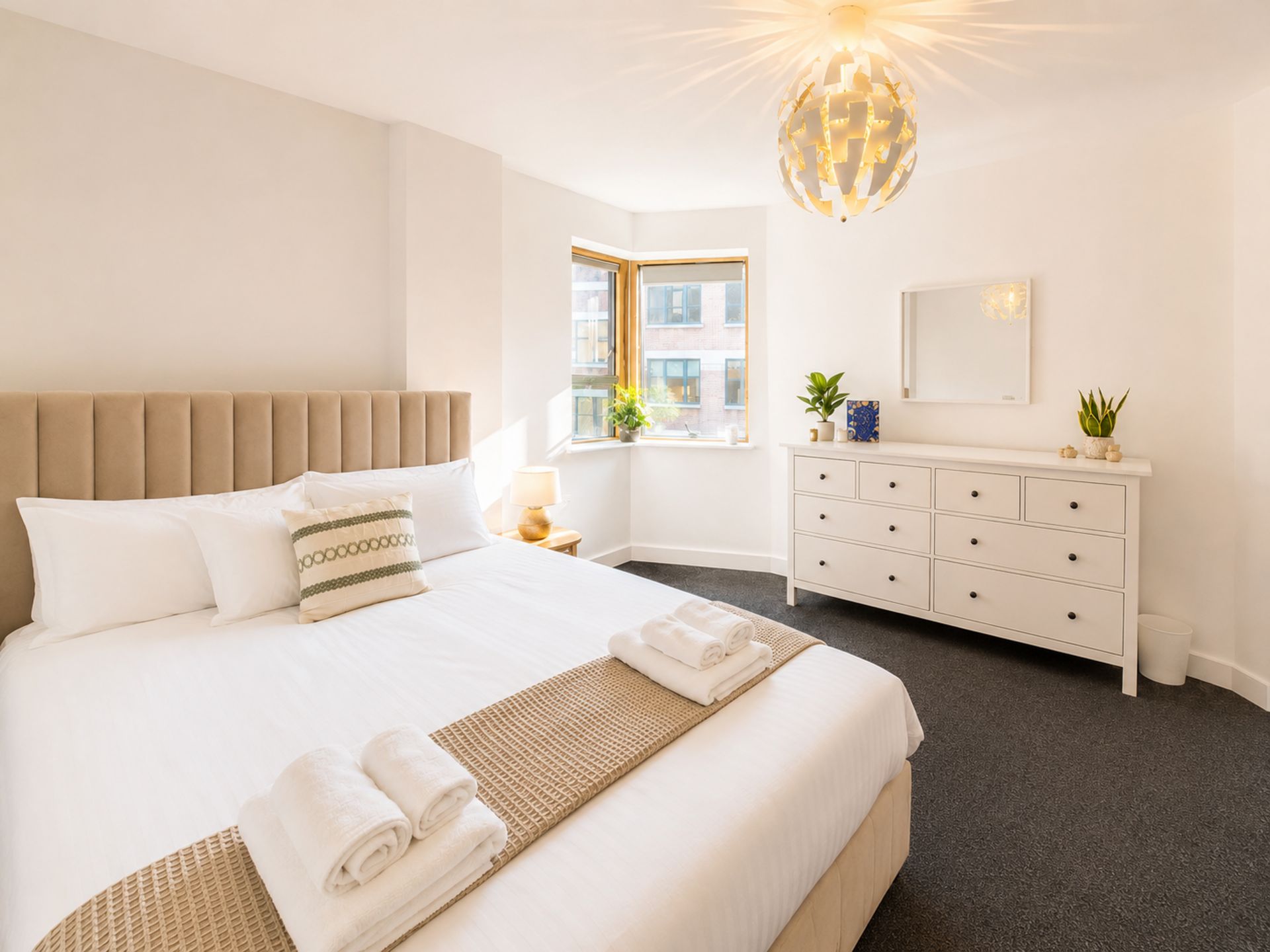 Une chambre d’hôtel avec un grand lit, une table de chevet, une commode, une fenêtre donnant sur des immeubles et un luminaire au plafond au design original.