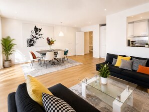 A modern living room with a black sofa, a glass coffee table, and a dining area with a white table and chairs. - Spacious 3BDR Haven in the Heart of East London (Greater London)