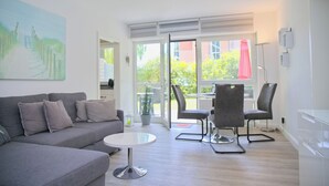 A modern living room with a grey sofa, a small round table, and two chairs. There is a large window with a view of greenery outside. - Sandkorn 6, cozy vacation rental in Grömitz with a terrace, garden, and 2 bedrooms (Grömitz)