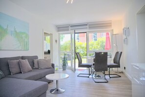 A modern living room with a grey sofa, a small round table, and two chairs. There is a large window with a view of greenery outside. - Sandkorn 6, cozy vacation rental in Grömitz with a terrace, garden, and 2 bedrooms (Grömitz)