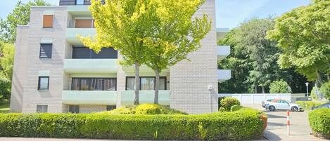 A modern multi-story residential building with balconies, a tree in the foreground, and a parking area with cars.