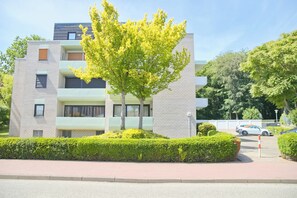A modern multi-story residential building with balconies, a tree in the foreground, and a parking area with cars. - Sandkorn 6, cozy vacation rental in Grömitz with a terrace, garden, and 2 bedrooms (Grömitz)