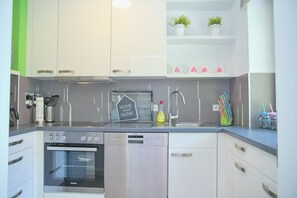 A modern kitchen with white cabinetry, a stainless steel dishwasher, a built-in oven, and a kitchen island with a sink. - Sandkorn 6, cozy vacation rental in Grömitz with a terrace, garden, and 2 bedrooms (Grömitz)