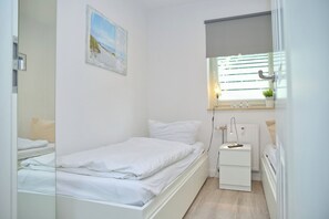 A small, clean room with a single bed, a bedside table with a lamp, and a window with blinds. - Sandkorn 6, cozy vacation rental in Grömitz with a terrace, garden, and 2 bedrooms (Grömitz)