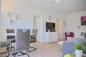 A modern living room with a dining area, a flat-screen TV mounted on the wall, and a small kitchen area. - Sandkorn 6, cozy vacation rental in Grömitz with a terrace, garden, and 2 bedrooms (Grömitz)