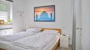 A bedroom with a bed, bedside tables, a nightstand, a window, and a wall-mounted picture. - Sandkorn 6, cozy vacation rental in Grömitz with a terrace, garden, and 2 bedrooms (Grömitz)