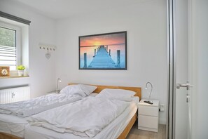 A bedroom with a bed, bedside tables, a nightstand, a window, and a wall-mounted picture. - Sandkorn 6, cozy vacation rental in Grömitz with a terrace, garden, and 2 bedrooms (Grömitz)