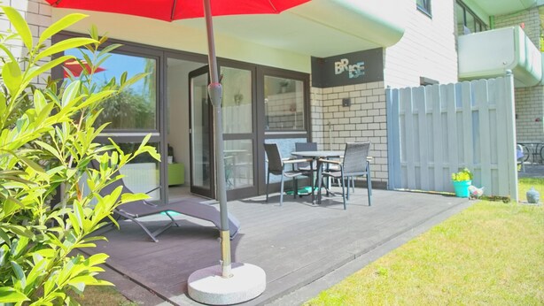 An outdoor patio with a red umbrella, a table and chairs, and a building with a sign that reads 'BRISE.' - Sandkorn 6, cozy vacation rental in Grömitz with a terrace, garden, and 2 bedrooms (Grömitz)