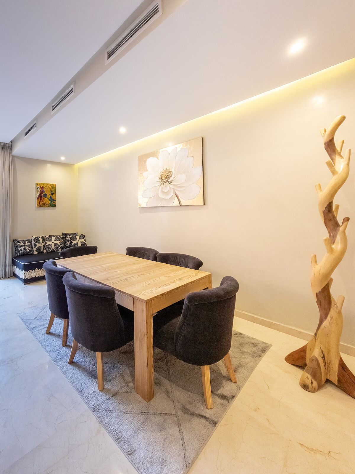Een eethoek met een houten tafel, zwarte stoelen, een bank met gekleurde kussens, een groot bloemen schilderij en een houten sculptuur.