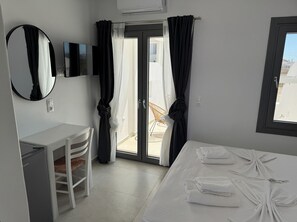 A hotel room with a bed, a desk, a chair, a TV, and a balcony with curtains. - Aitheria Suites Ios (Ios)