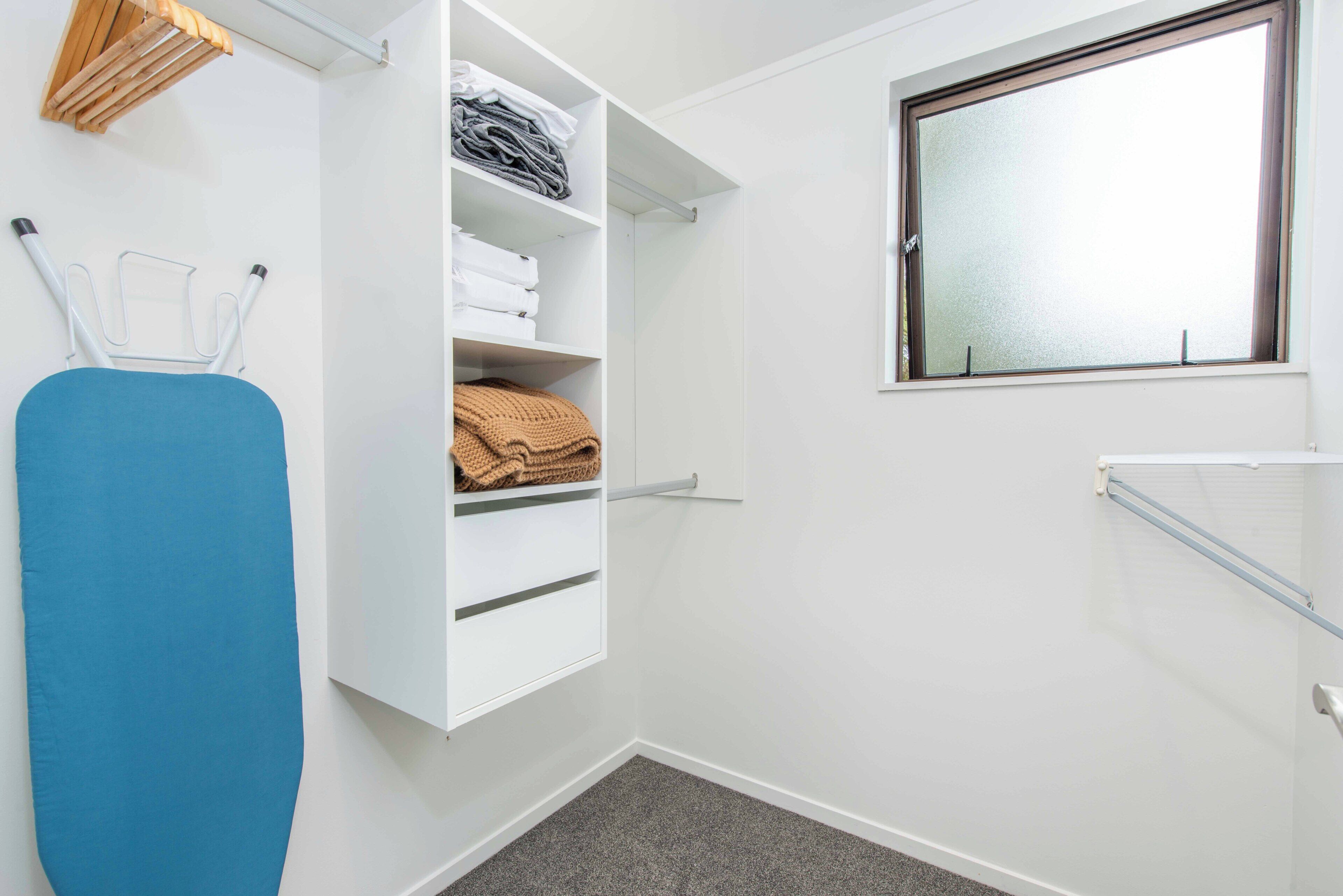 A neatly organized laundry room with a blue ironing board, shelves with folded clothes, and a window with a view.