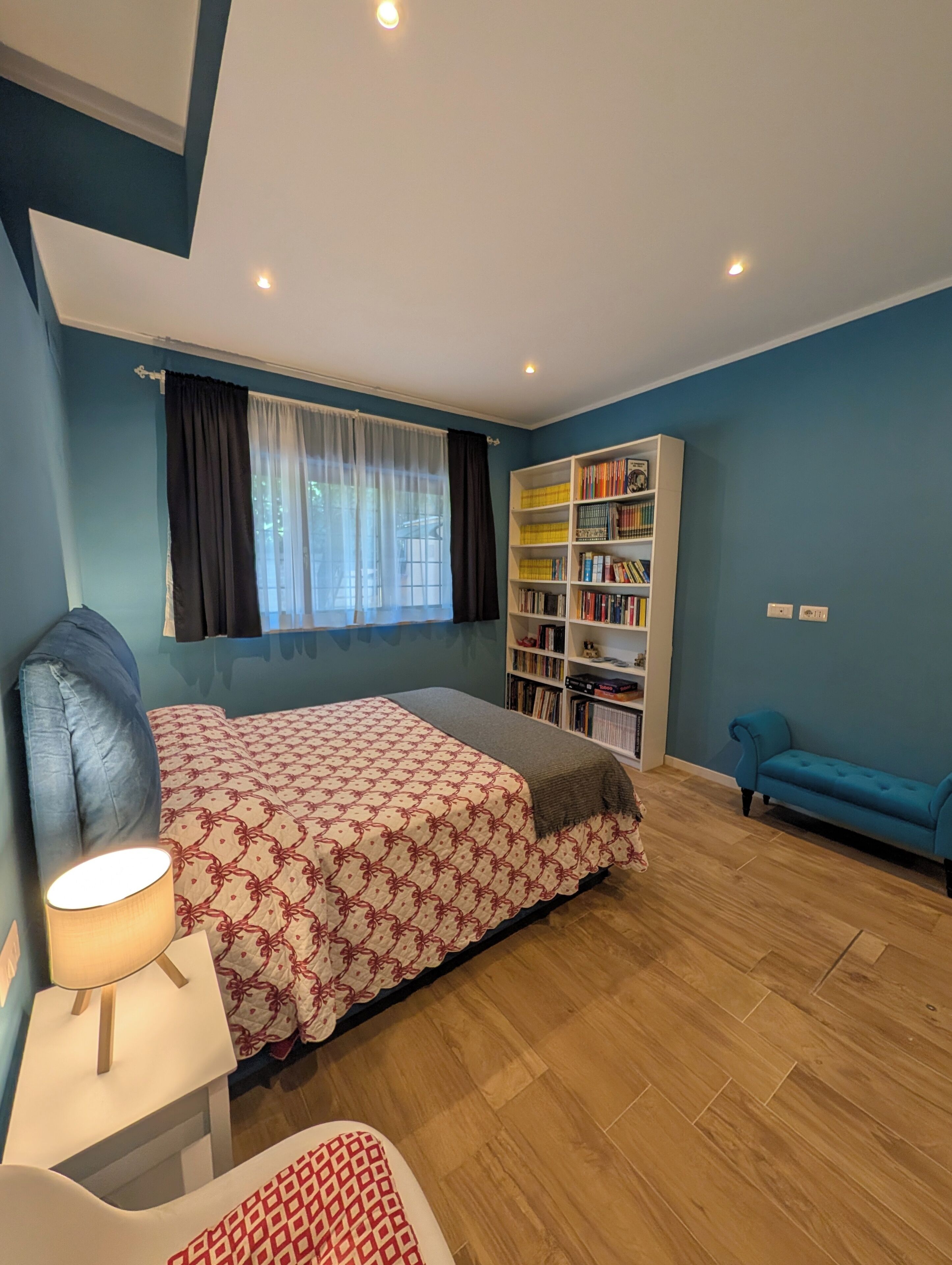 A bedroom with a bed, a bedside table with a lamp, a bookshelf, and a blue sofa.