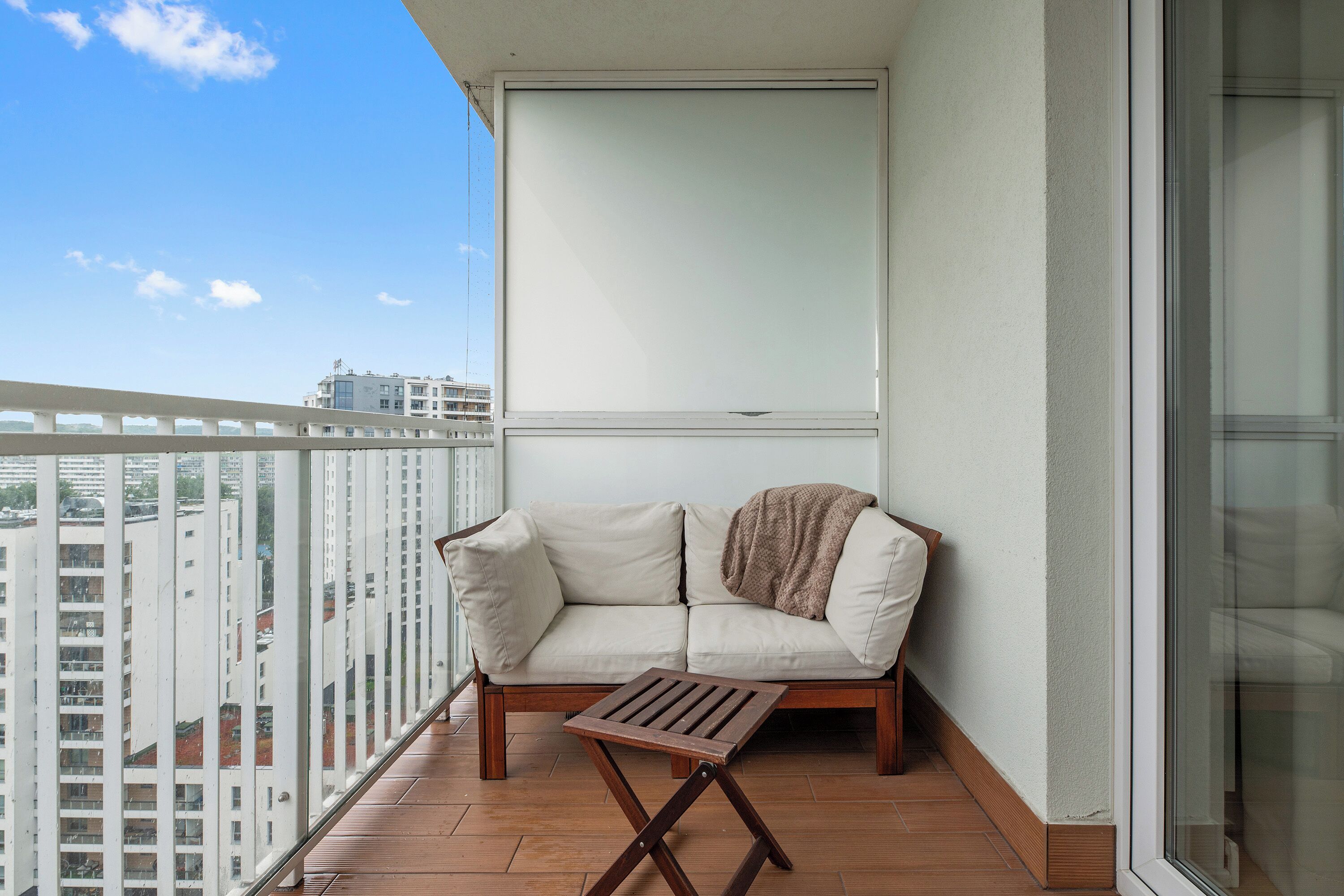 A balcony with a sofa and a small table, a view of buildings, and a clear sky.