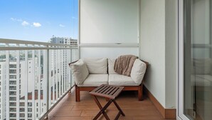 Uma varanda com um sofá e uma mesa pequena, vista para prédios e céu claro.