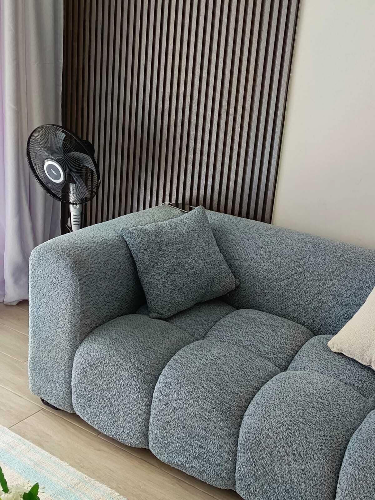 A sectional sofa with a textured fabric, a standing fan, and a vertical slatted wall in the background.