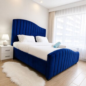 A modern bedroom with a blue upholstered bed, white bedding, a white fluffy rug, a bedside table with a lamp, and a large window with sheer curtains. - Modern 2BR Near JKIA & SGR with WiFi, Gym & Secure Parking (Nairobi)