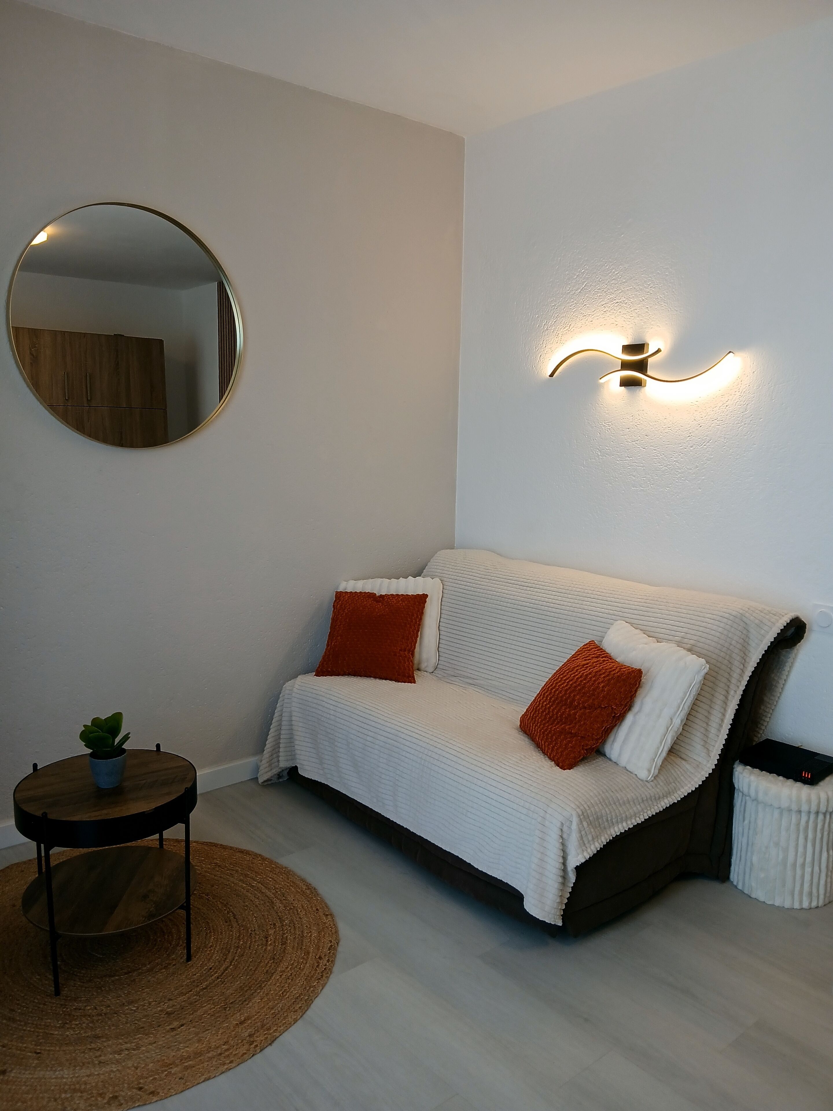 Uma sala de estar moderna com um sofá, um espelho redondo, uma luminária fixada na parede e uma pequena mesa redonda com uma planta.