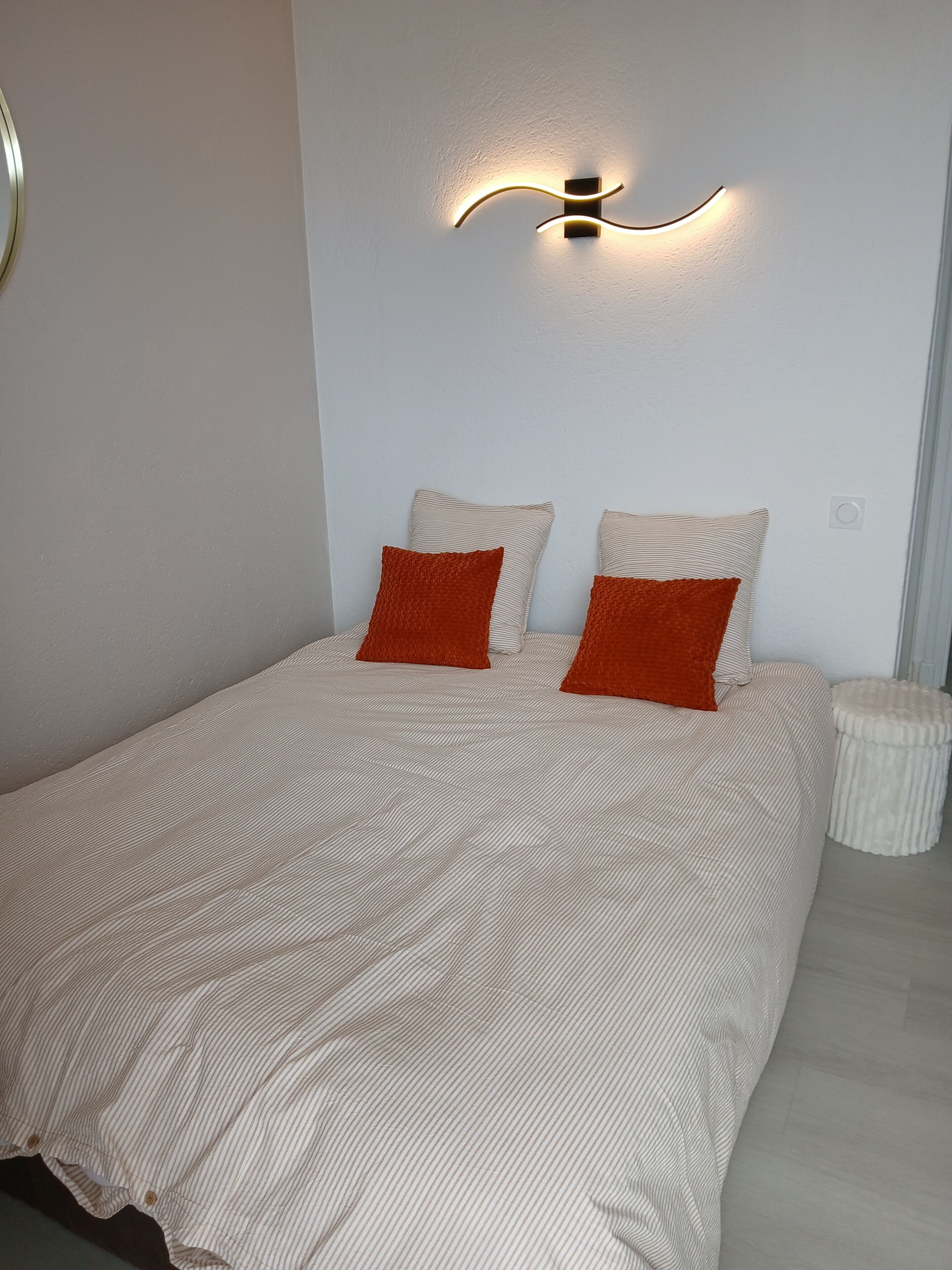 Una cama bien hecha con ropa de cama a rayas y dos almohadas, un aplique en la pared y un taburete pequeño y redondo.