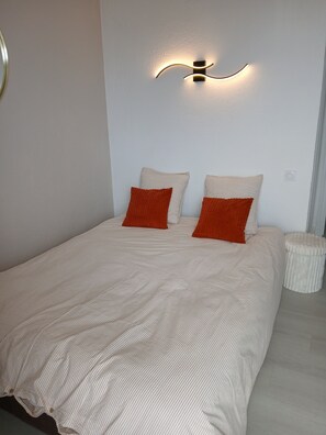 A neatly made bed with striped bedding and two pillows, a wall-mounted light fixture, and a small round stool.