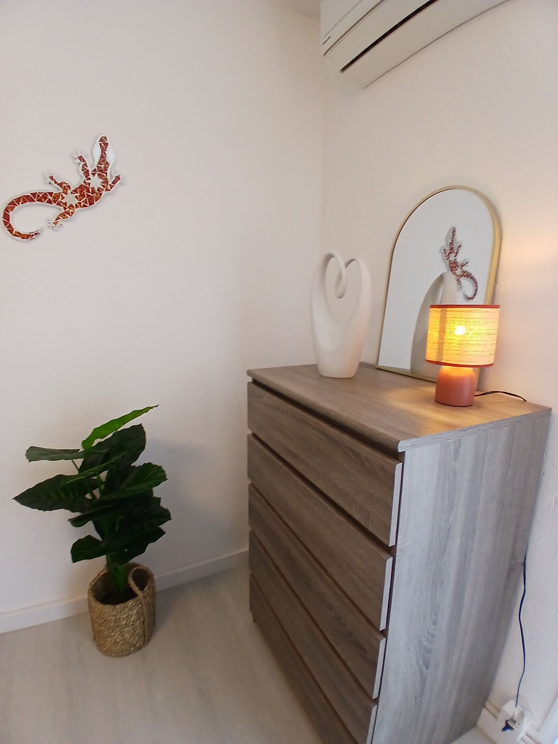 A room with a wooden dresser, a potted plant, a wall-mounted mirror, and a decorative wall piece.