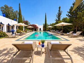 Un área de piscina con sillones de descanso y sombrillas, rodeada de vegetación y un cielo despejado.