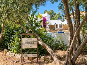 A wooden sign with 'UNE PARTIE DE PÉTANQUE' and a number sequence, surrounded by trees and a pool area. - Palais Mogaya (Essaouira)