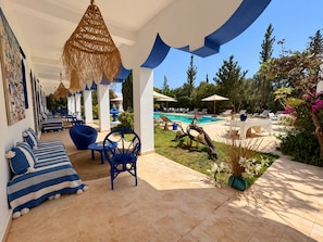 A covered outdoor seating area with blue furniture, a pool, and a garden view. - Palais Mogaya (Essaouira)